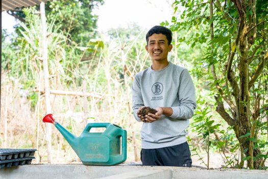 A young man holds a plant and dirt and smiles at the camera.
