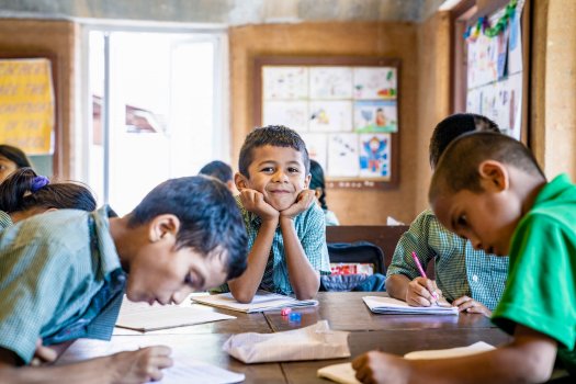 Children write at a group of desks, while one boy smiles at the camera.