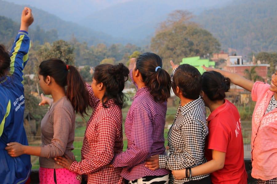 A group of people stands outdoors, arms linked, looking and pointing towards a scenic landscape with hills and trees in the background. They are dressed in casual, colorful clothing. The mood appears joyful and relaxed.