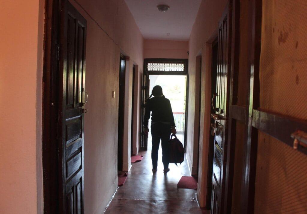 Silhouette of a person walking down a dimly lit hallway, carrying a bag. There are doors on both sides, and light is visible at the end of the corridor through an open door.