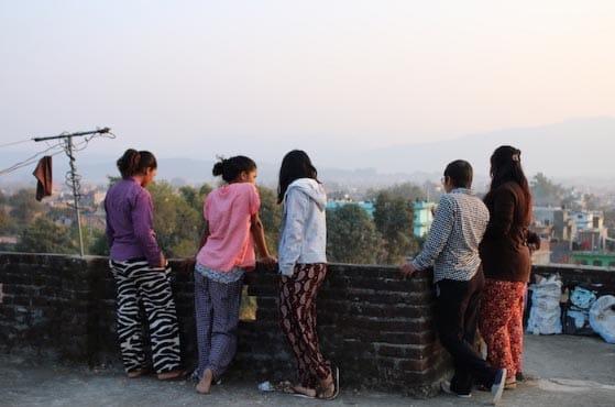 A group of five people stand on a rooftop, leaning against a brick wall and looking at the cityscape during sunset. They are dressed in casual, colorful clothing, and the background shows trees and distant hills.