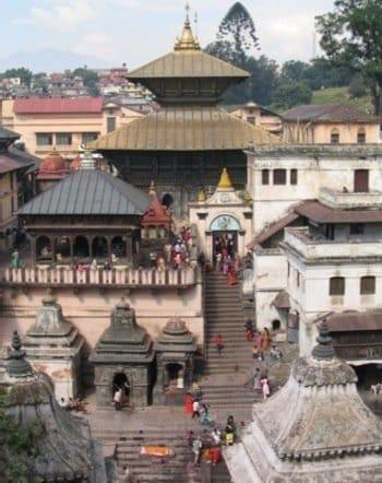 A bustling scene at the Pashupatinath Temple in Kathmandu, Nepal, showcasing its intricate architecture. People are gathered around, with several shrines and a prominent golden-roofed temple visible. Trees and city buildings are in the background.