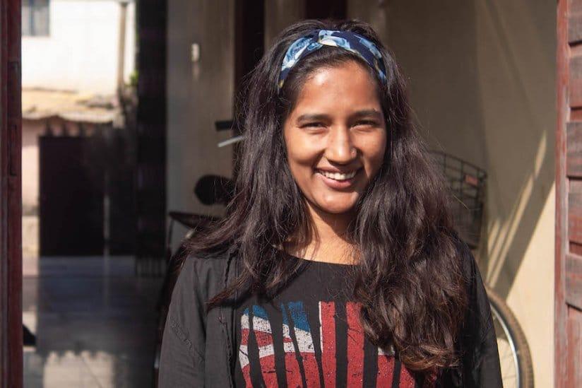 A person with long hair stands smiling in a sunlit corridor. They wear a blue and white headband and a dark jacket over a shirt with a Union Jack design. A bicycle and open doorway are visible in the background.