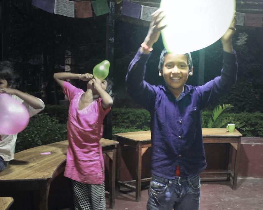 Children joyfully inflating balloons outside, with one child in a pink outfit and another in a blue shirt smiling brightly. The scene is set in a dimly lit area surrounded by greenery.