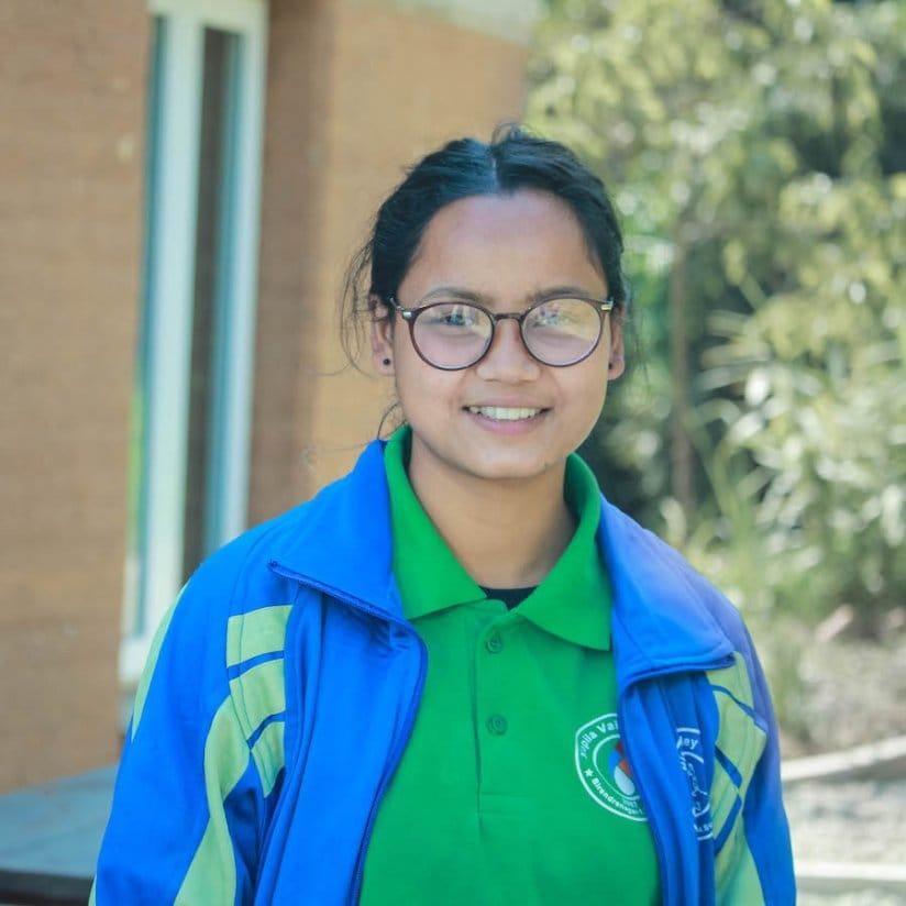 A young person wearing glasses, a green shirt, and a blue jacket with yellow stripes stands outside smiling. The background features greenery and a brick building.