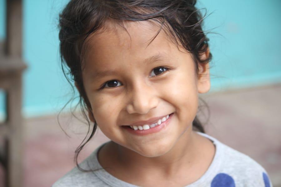 Smiling child with long dark hair, wearing a gray shirt with blue spots, against a light blue background.