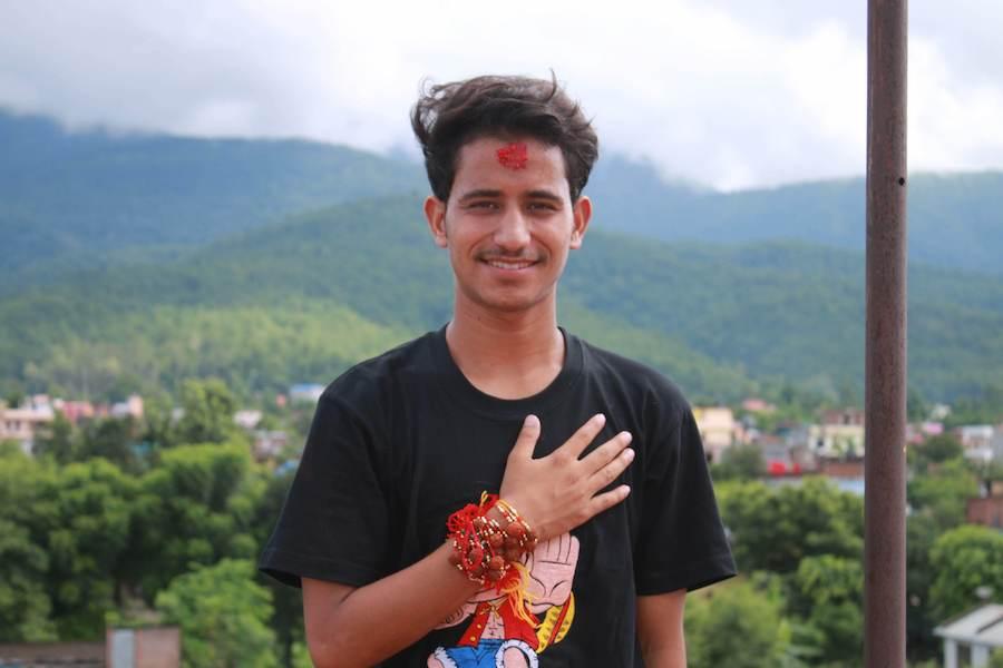 A young man wearing a black t-shirt and a red tikka on his forehead stands outdoors with a mountainous backdrop. He places his right hand on his chest, adorned with red and yellow threads and bracelets. The background features greenery and cloudy skies.