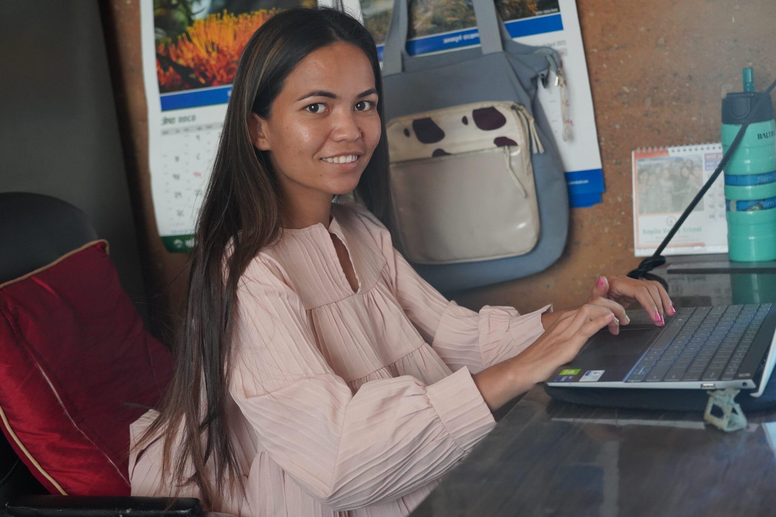 A woman with long hair and a pink blouse is sitting at a desk using a laptop. She is smiling while looking at the camera. Behind her are hanging calendars, a bag, and a large water bottle on the desk.