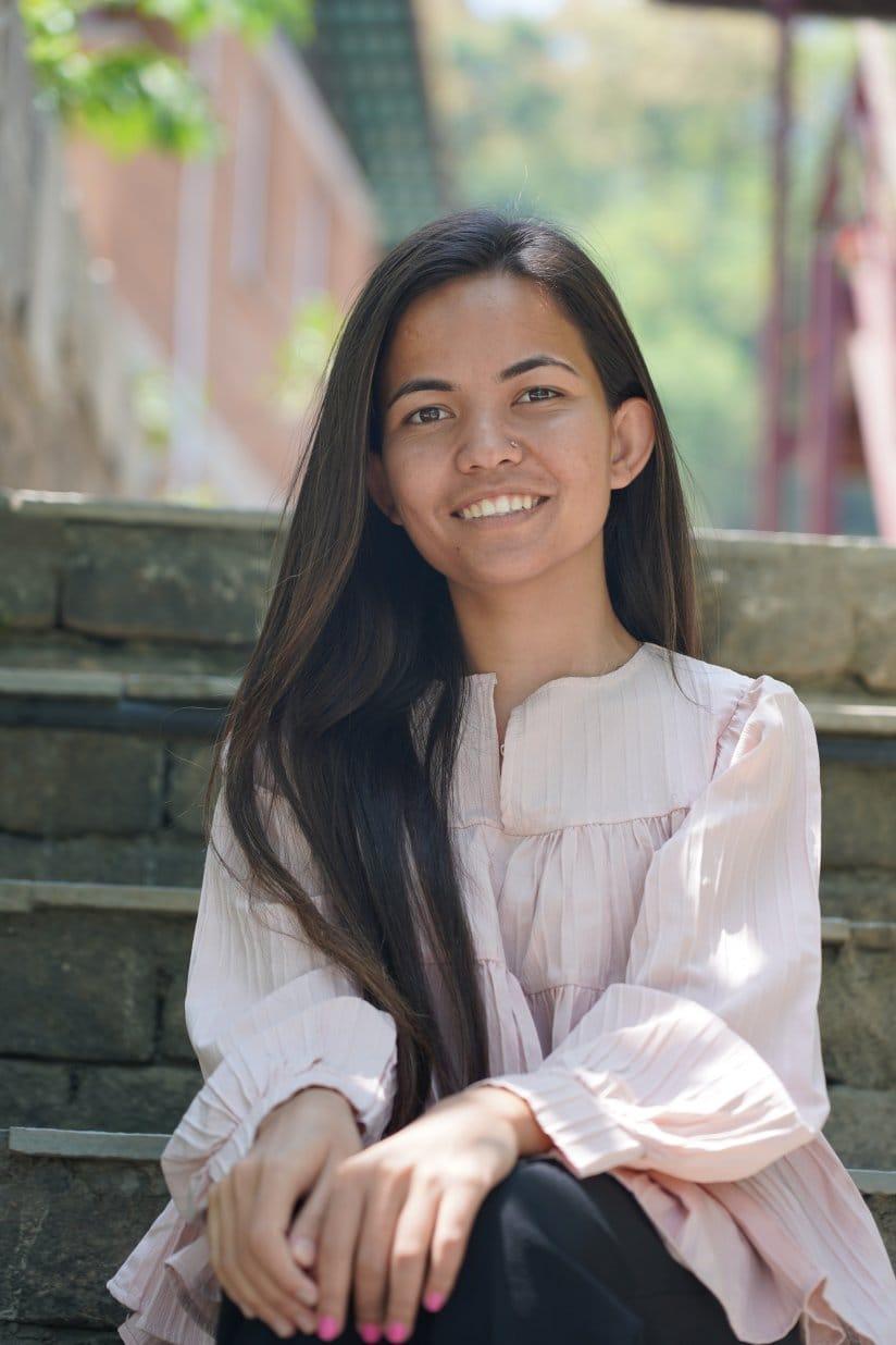 A woman with long dark hair sits on stone steps outdoors, smiling at the camera. She is wearing a light pink blouse, and there is greenery and a building in the background.