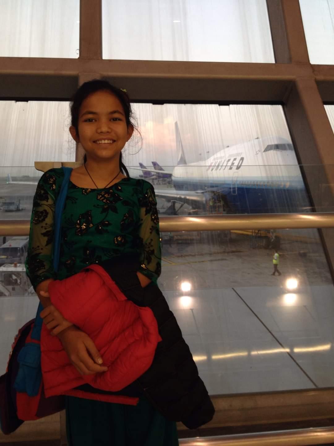 A young girl smiles at the camera while standing inside an airport terminal. She holds a red jacket and is wearing a green dress. Behind her, through large windows, a United Airlines plane is visible parked on the tarmac.