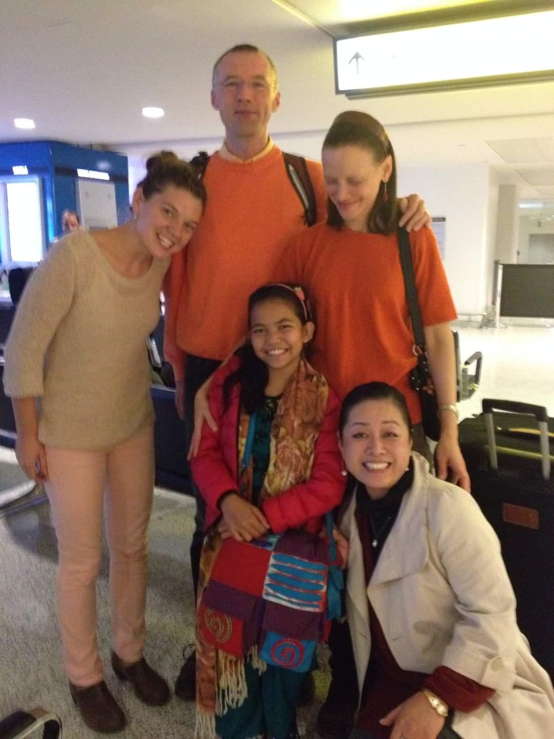 A group of five people smile for a photo at an airport. Two adults stand behind, one wearing an orange sweater. Three others, including a young girl in colorful attire, are in front. Luggage and signage are visible in the background.