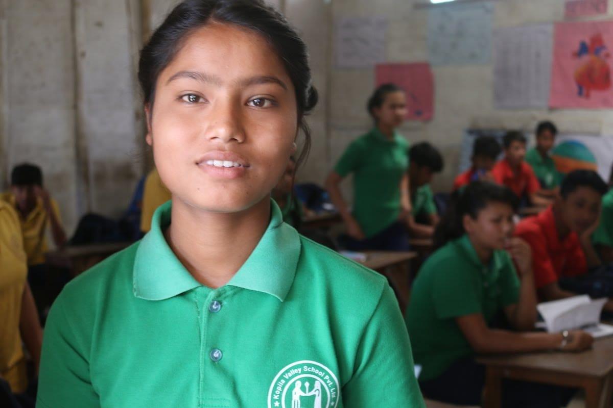 A young student in a green polo shirt stands in the foreground of a classroom, with other students seated at desks in the background. The classroom walls feature colorful educational posters.