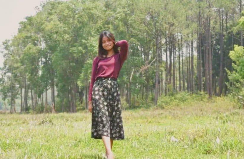 A person stands in a grassy field, smiling with one hand behind their head. They wear a maroon top and a black floral skirt. A row of tall trees is visible in the background under a clear sky.