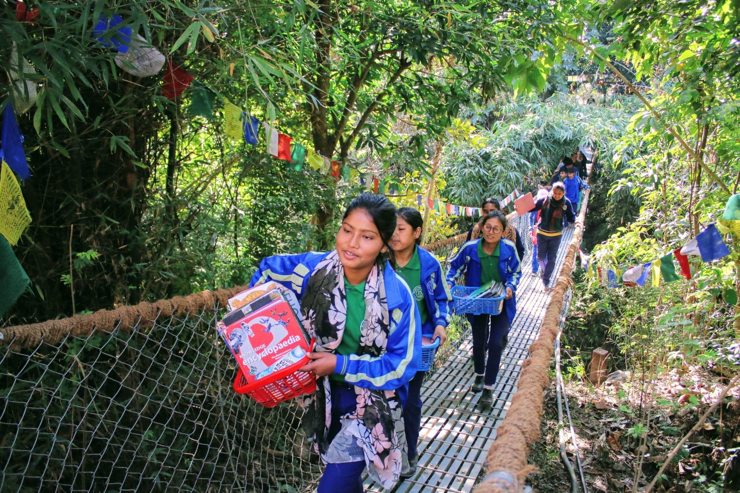 A group of people carrying boxes and supplies walk across a narrow, elevated wooden bridge in a lush, green forest. Colorful prayer flags hang along the bridge, fluttering in the breeze.