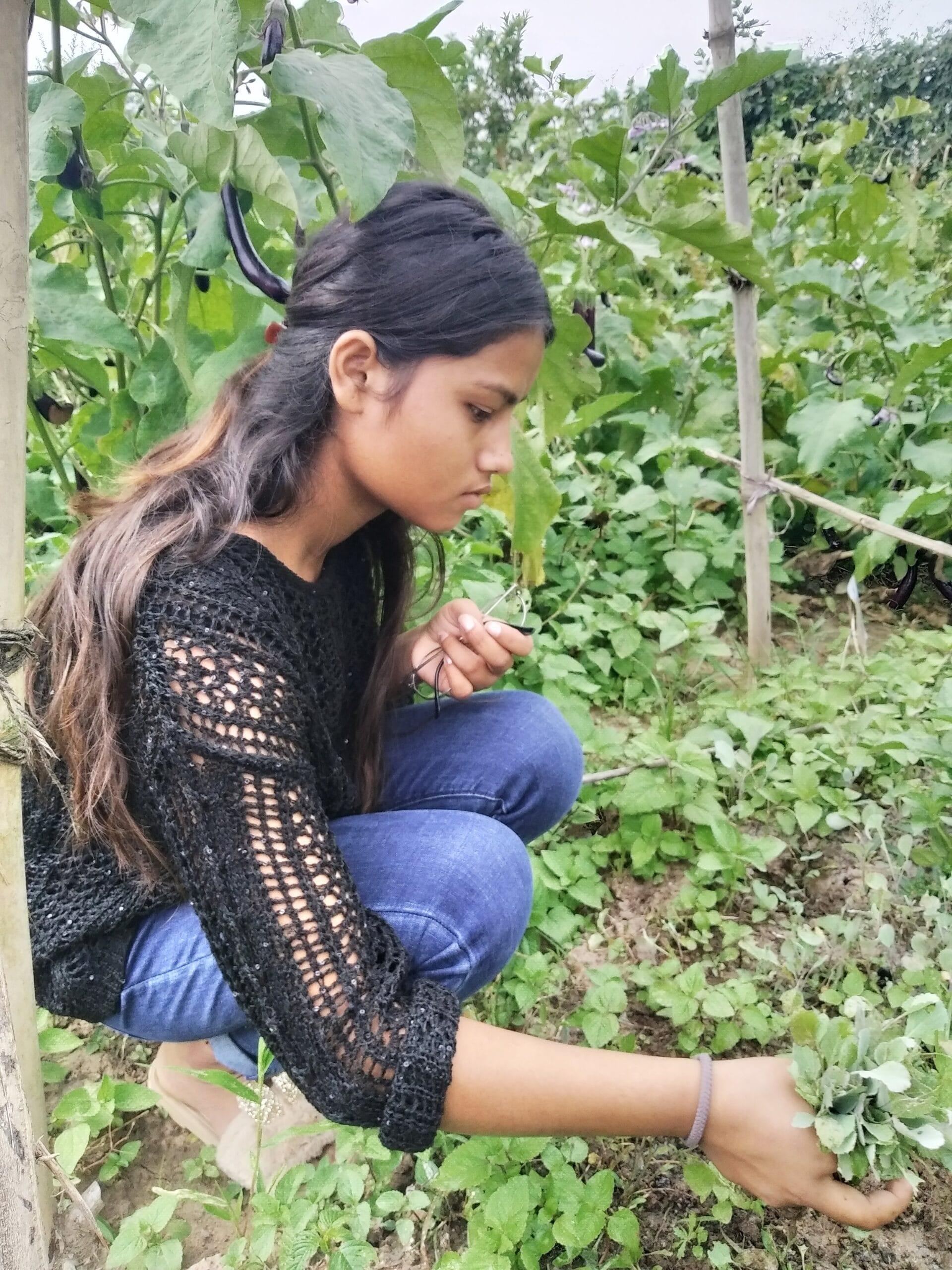 A young woman crouches in a garden, picking herbs or vegetables. She has long dark hair and is wearing a black crochet top and blue jeans. Green plants surround her in a lush outdoor setting.