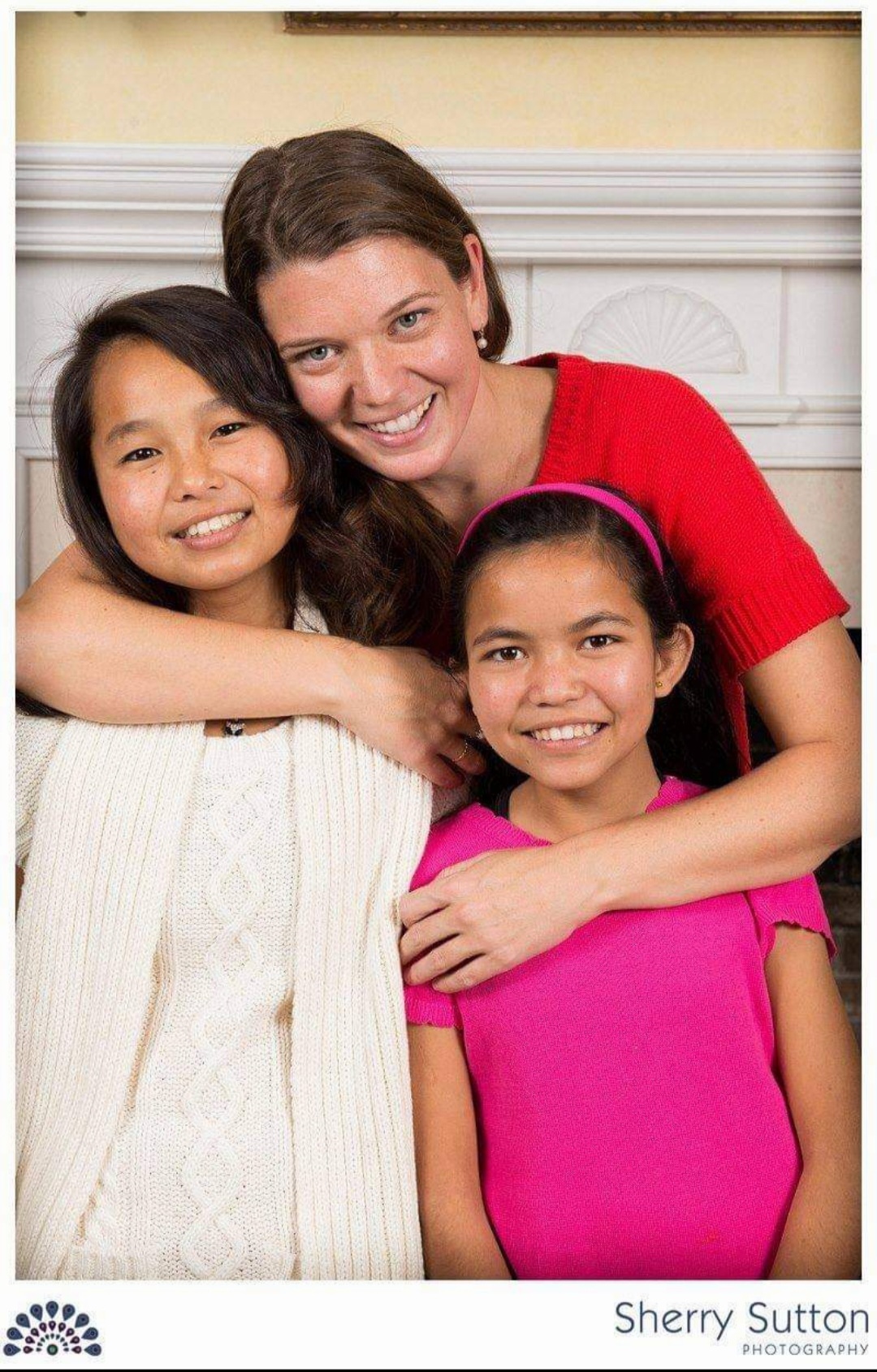 A woman wearing a red sweater smiles and embraces two young girls. The girl on the left wears a cream sweater, and the girl on the right wears a pink shirt with a matching headband. They are standing in front of a white backdrop indoors.