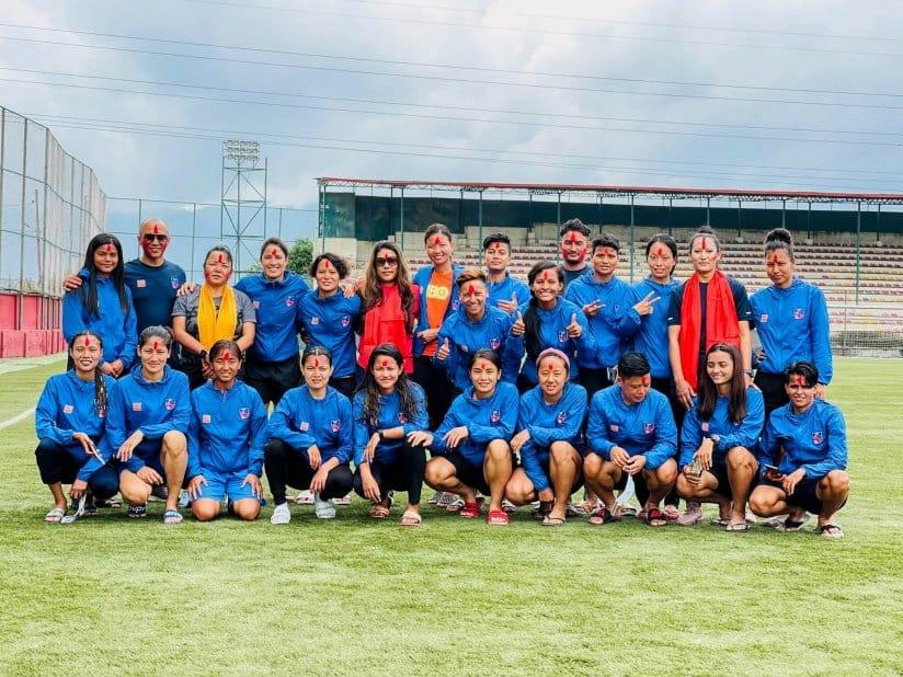 A group of soccer players in blue jerseys pose together on a field. Some are standing, while others are kneeling. They seem cheerful, with a stadium and cloudy sky in the background.