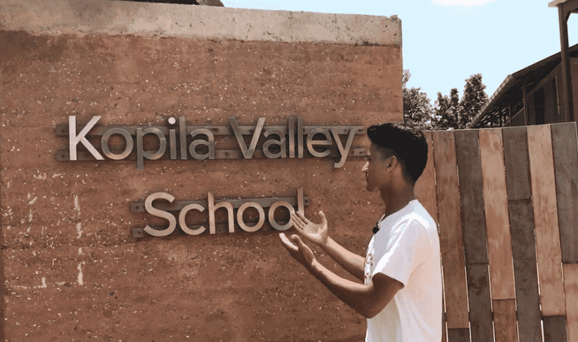 A person standing in front of a wall with the words "Kopila Valley School." They have short dark hair and are facing the wall, gesturing with their hands. The setting appears to be outdoors with clear skies and trees in the background.