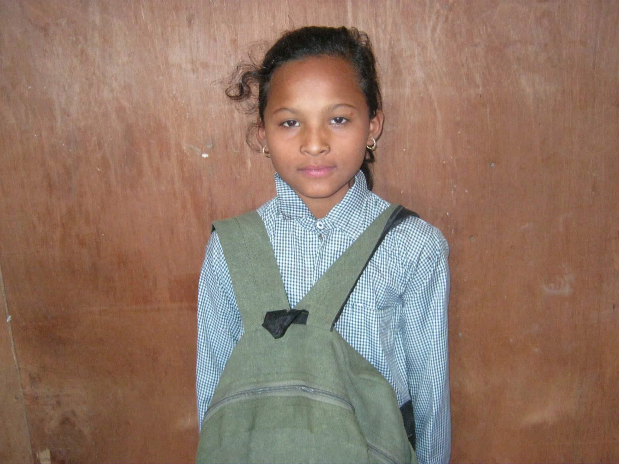 A young girl stands against a wooden background, wearing a checkered shirt and a green backpack. She has curly hair and small hoop earrings, looking directly at the camera with a neutral expression.