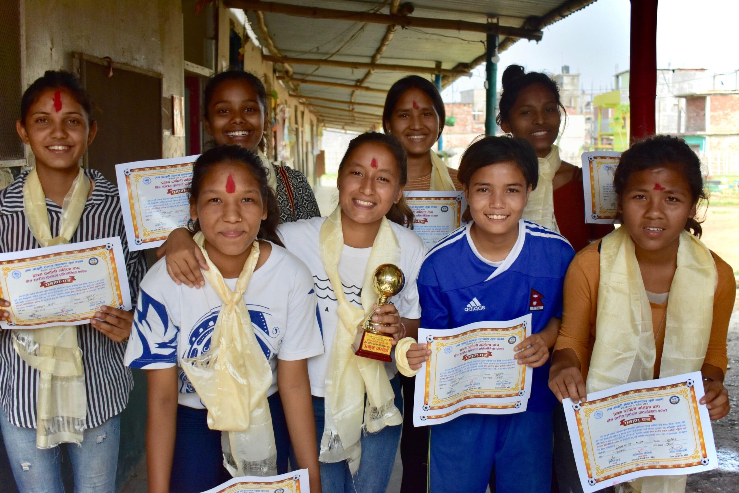 A group of eight young people standing together, holding certificates and a trophy. They are smiling and wearing traditional yellow scarves and red tikas on their foreheads, standing in a covered outdoor area.