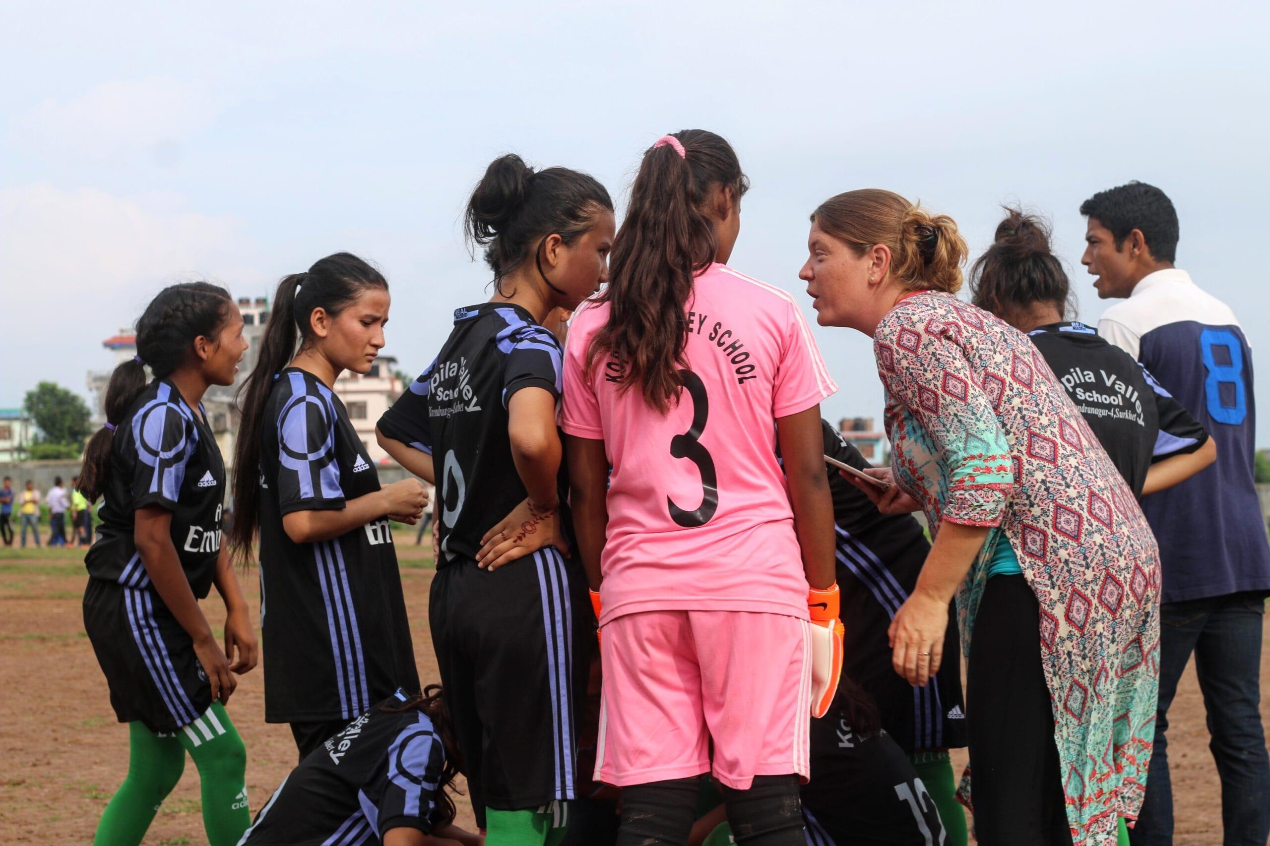 A group of young female soccer players in black jerseys are gathered on a field, listening to a coach in a patterned dress. One player in a pink jersey stands out, and a few onlookers are in the background. The scene conveys teamwork and guidance.