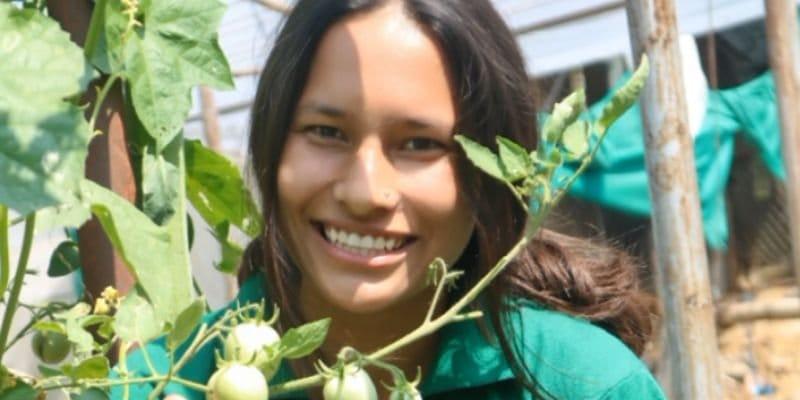 A woman smiling and leaning near green plants with small tomatoes, dressed in a green shirt. There are wooden stakes and a blurred background, suggesting a garden or greenhouse setting.