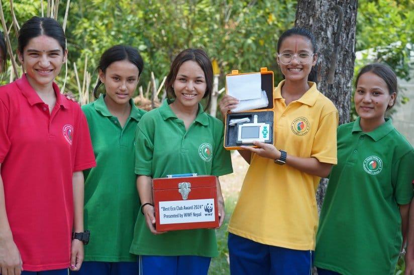 Five smiling people stand outdoors wearing colored shirts. Two hold orange cases with a sign that reads "Precitas Club Award 2022 Preserved by World Nepal." They pose proudly with the equipment, surrounded by greenery.