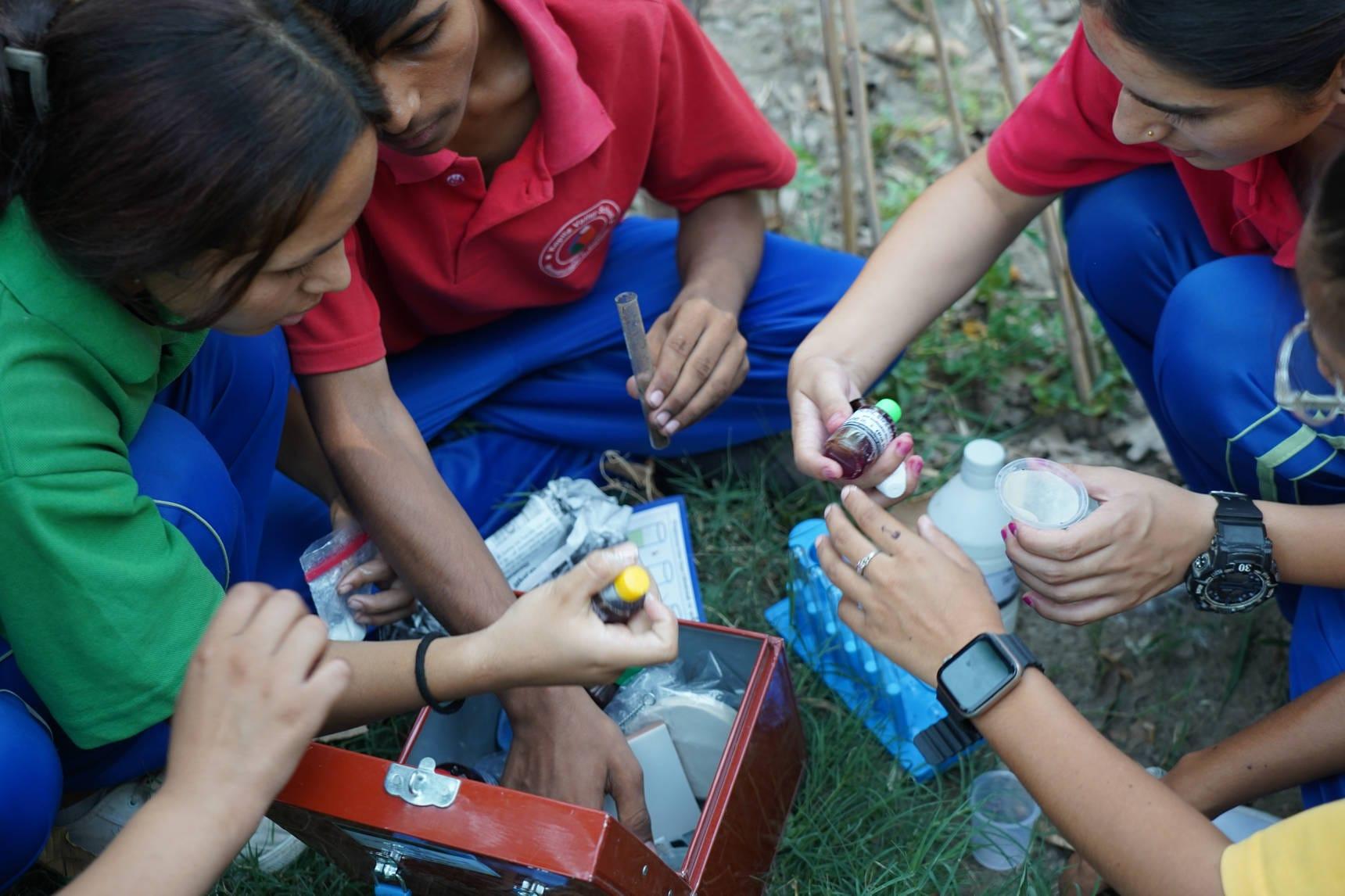 A group of people sitting outdoors, engaged in a hands-on activity with scientific equipment. They are wearing colorful shirts and appear focused on their work, examining items like a bottle and a test tube from a kit.