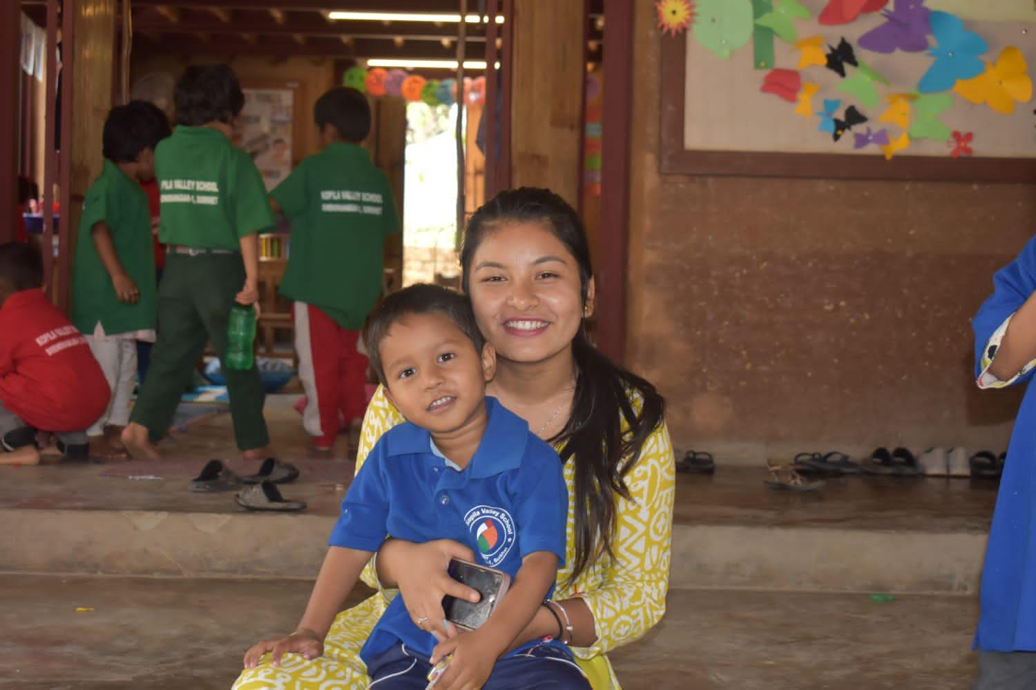 A woman smiles while holding a young boy wearing a blue shirt. They are indoors, with children dressed in colorful uniforms playing in the background. Bright artwork decorates the walls.