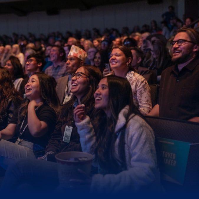 A diverse audience is seated in a dimly lit theater, smiling and laughing while watching a performance or presentation. Some people are holding popcorn, and the atmosphere is lively and engaged.
