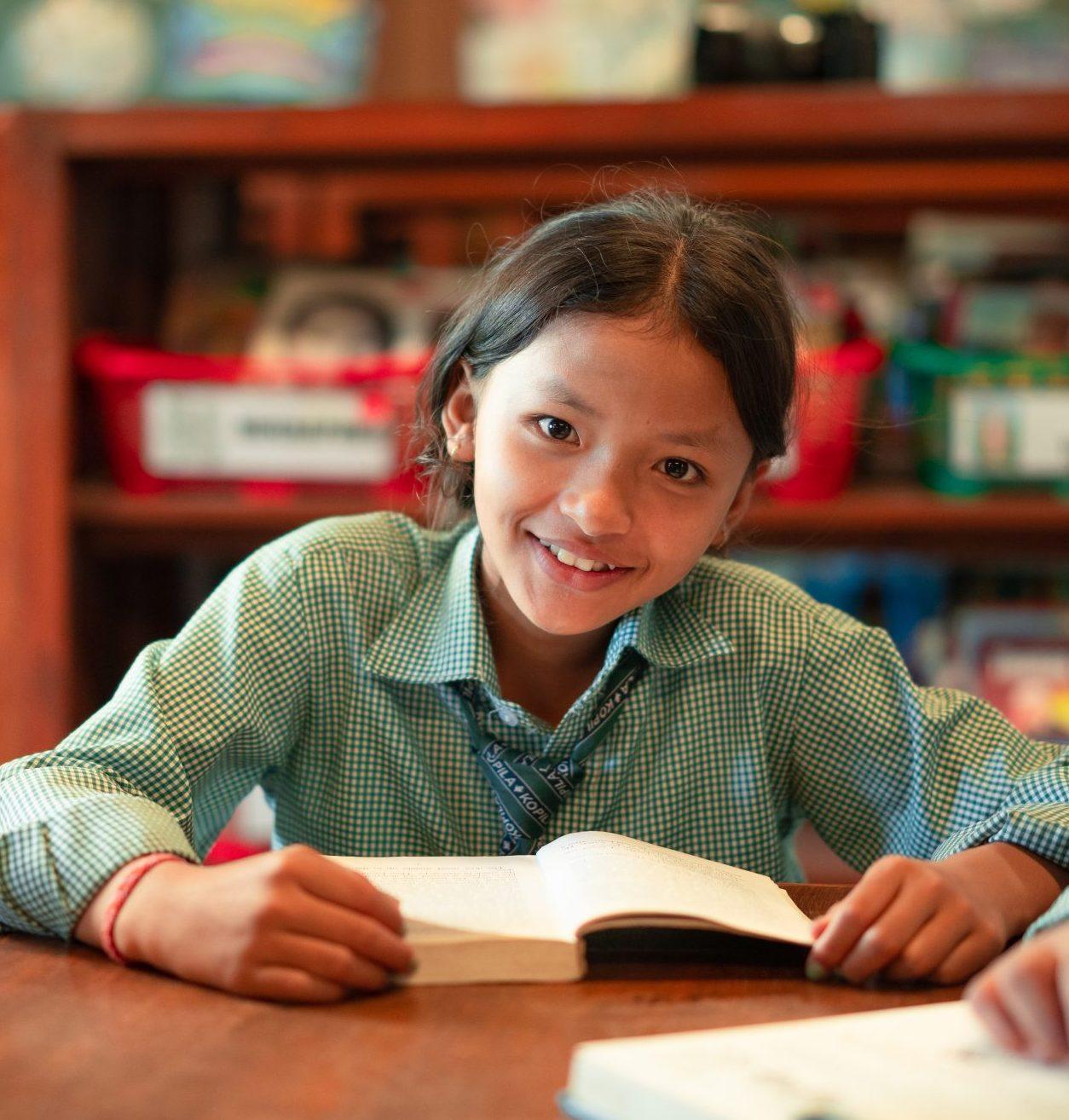 A girl in school uniforms sit at a wooden table, reading books. She looks directly at the camera, smiling. A bookshelf with colorful items is in the background.