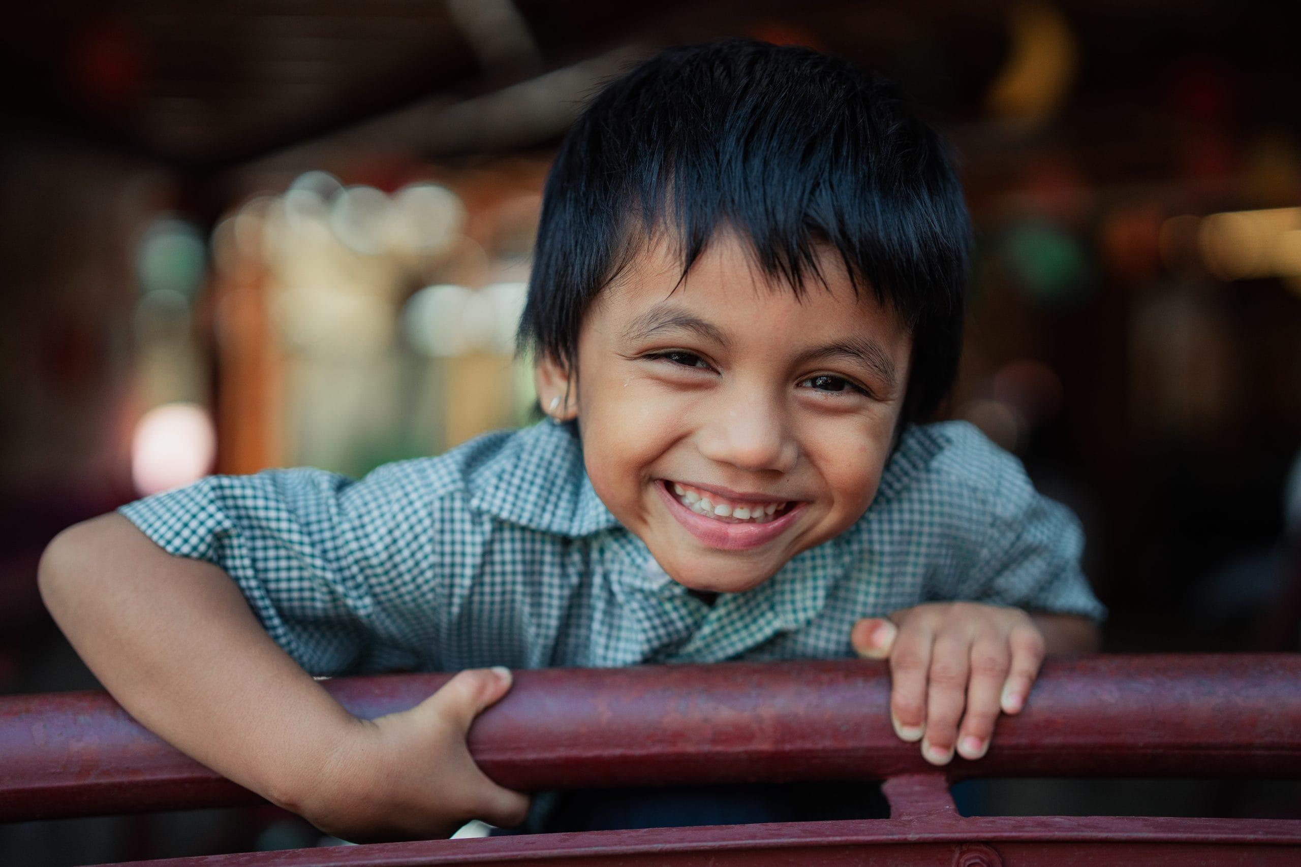 A young boy with black hair and a checkered shirt leans on a red railing, smiling brightly at the camera. The background is slightly blurred, suggesting an indoor setting with soft lighting.