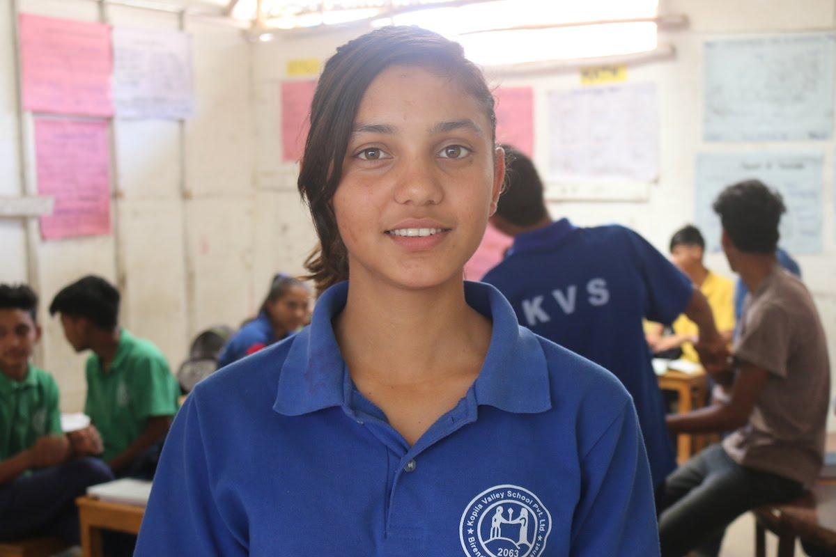 A young person in a blue shirt stands smiling in a classroom setting, with a group of students in the background engaging in activities. Classroom walls have colorful posters.