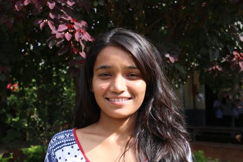 A person with long dark hair smiles in front of purple and green foliage. They are wearing a white top with blue and red patterns. The sunlight is casting soft shadows.