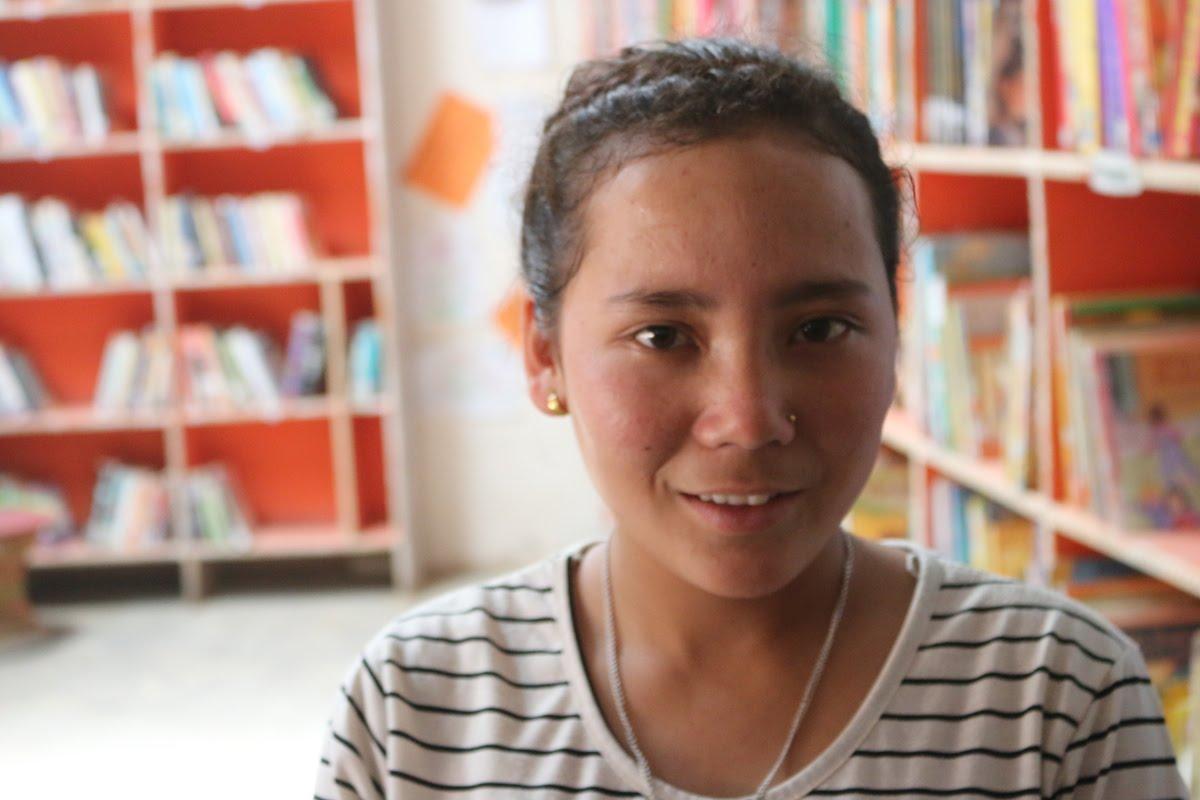 A person wearing a striped shirt smiles in the foreground. Behind them, bookshelves filled with various books are visible, creating a library setting.