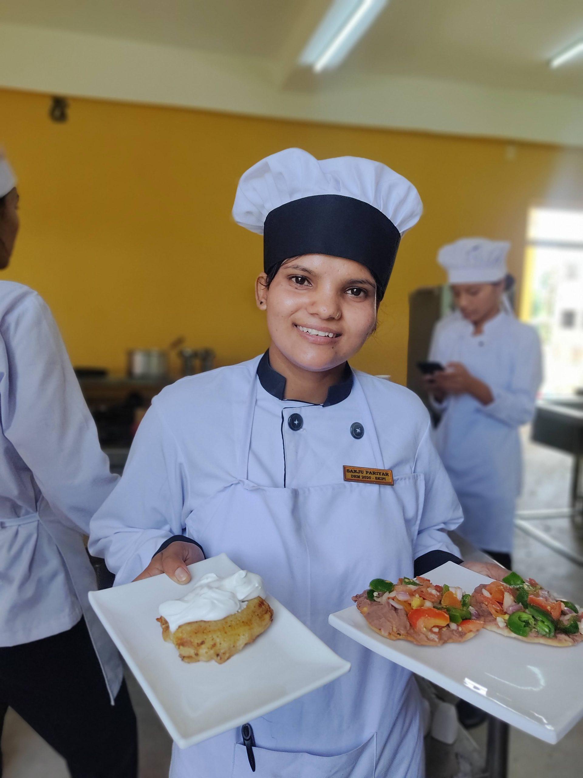 A chef in a white uniform and black hat smiles while holding two plates of food. One plate has a dessert topped with cream, and the other features a dish with vegetables and meat. Other chefs are seen working in the background.