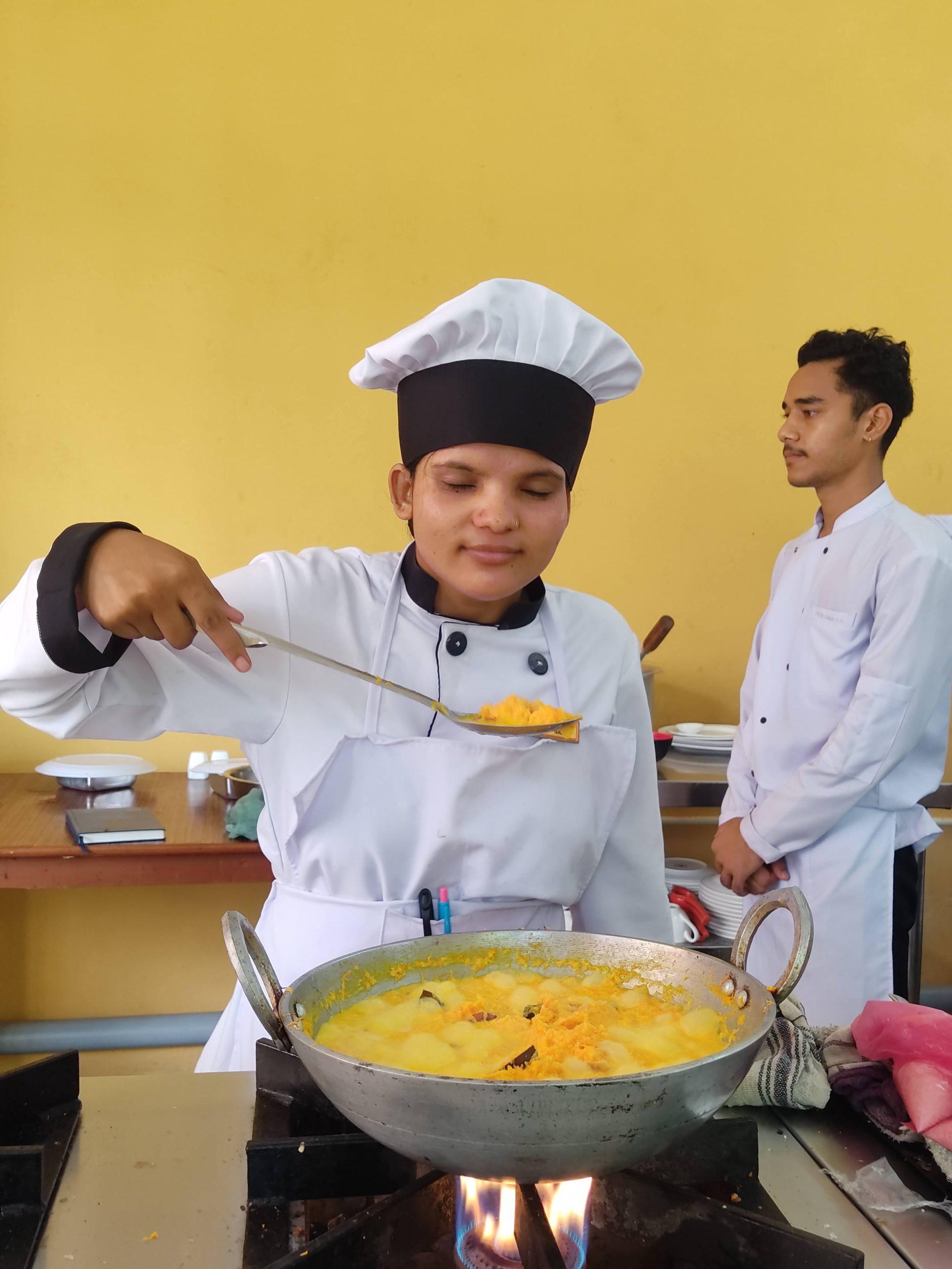 A chef wearing a white uniform and black hat stirs a pan of food on a stove. A yellow wall is in the background, and another person in a white chef's jacket stands nearby.