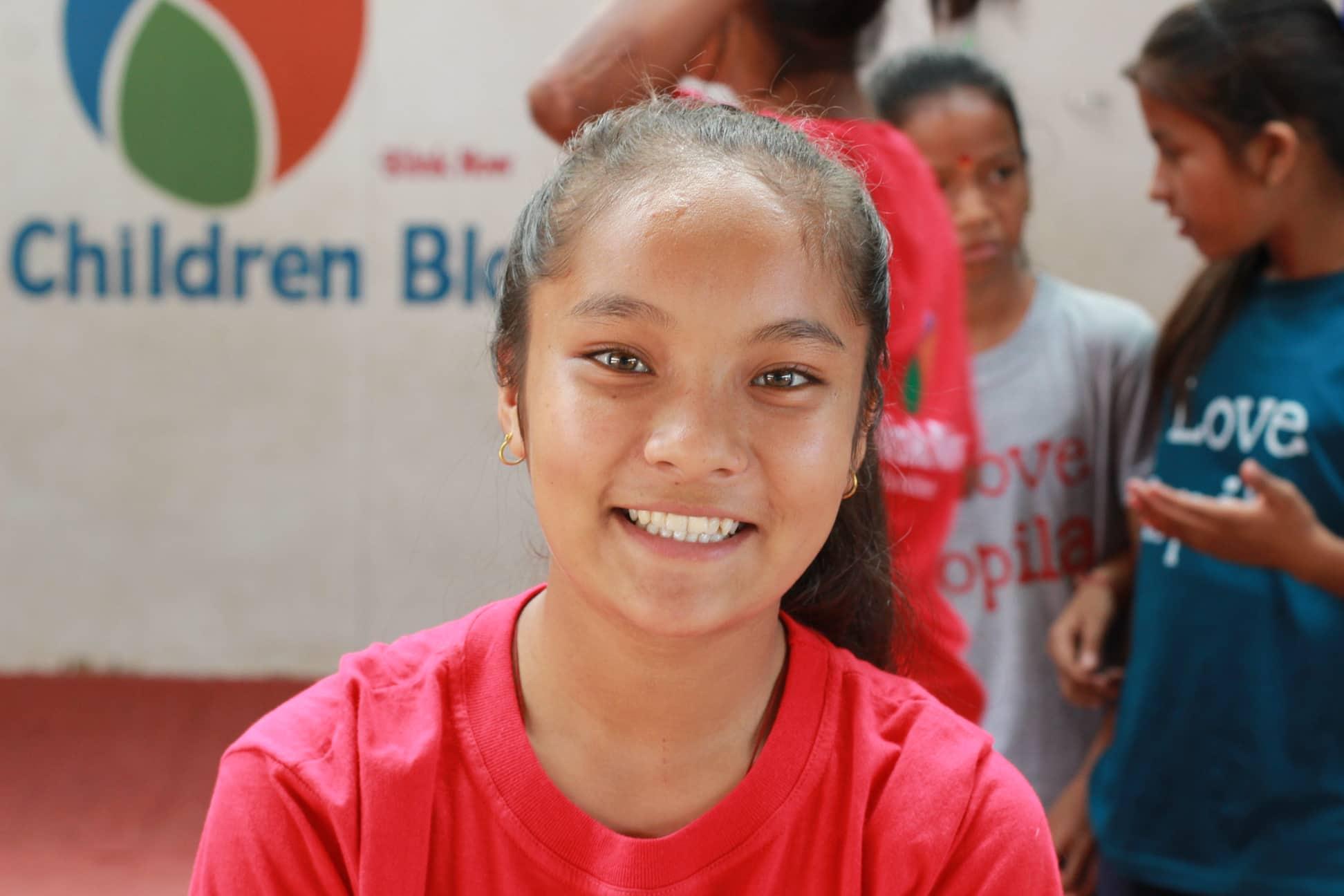 A smiling girl in a red shirt stands in front of a blurred background with other children. She is indoors, and a colorful wall with the partially visible word "Children" is behind her.