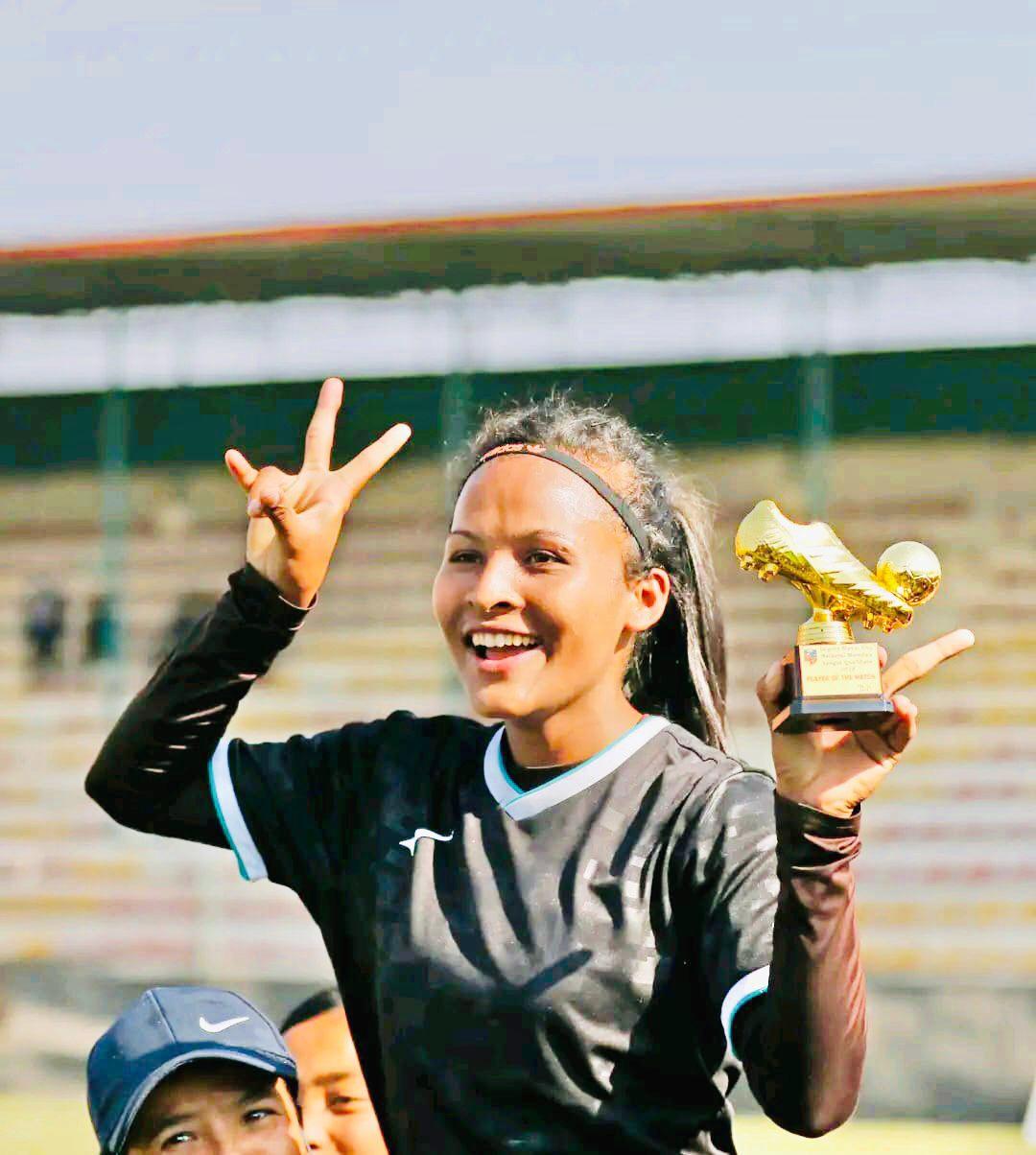 A soccer player in a black jersey holds up a gold trophy shaped like a shoe, smiling enthusiastically. She is standing on a field with a blurred stadium background. Other people, slightly obscured, are around her.