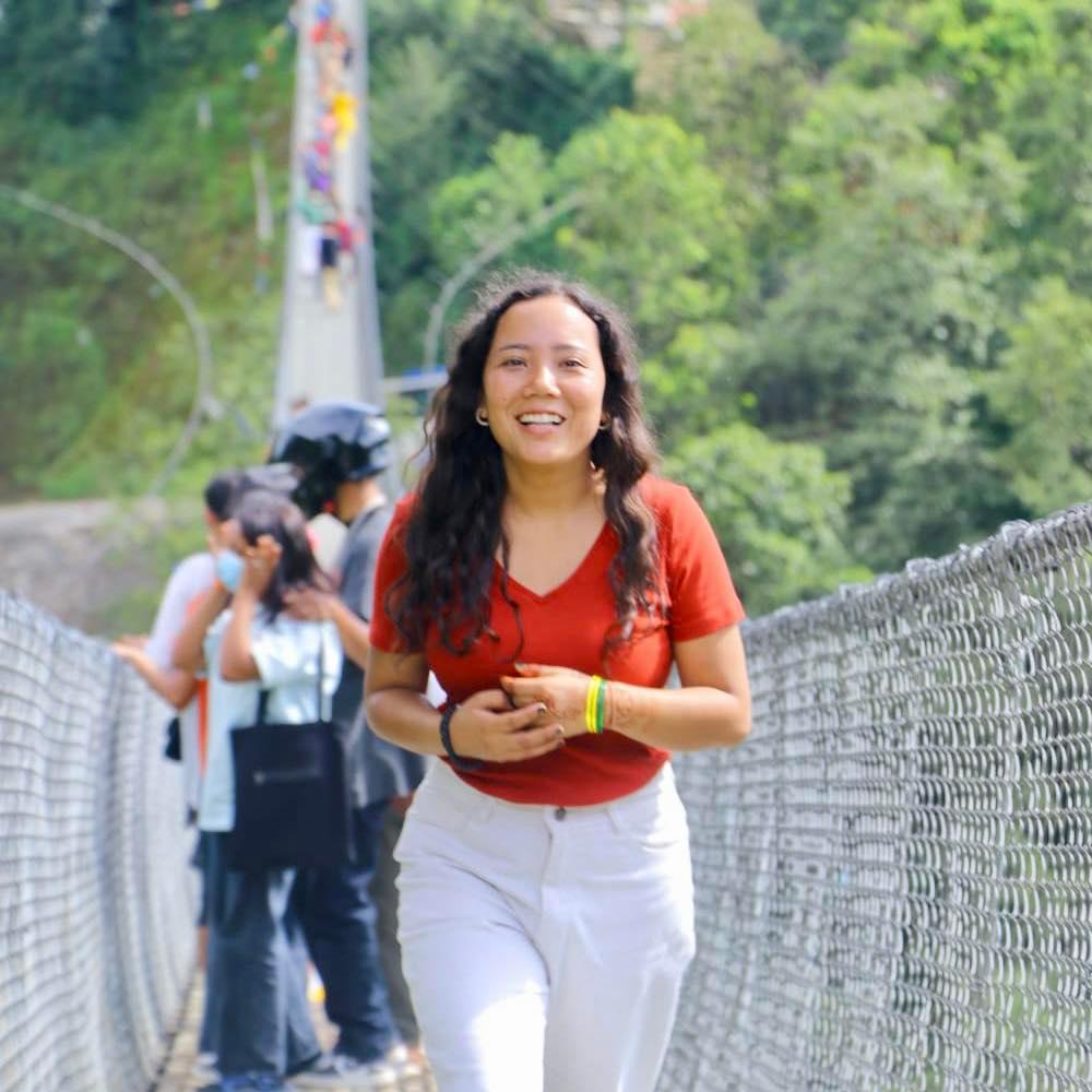 A woman in a red top and white pants walks toward the camera on a suspension bridge. She is smiling, and there are people in the background also crossing the bridge. Lush green foliage surrounds the area.