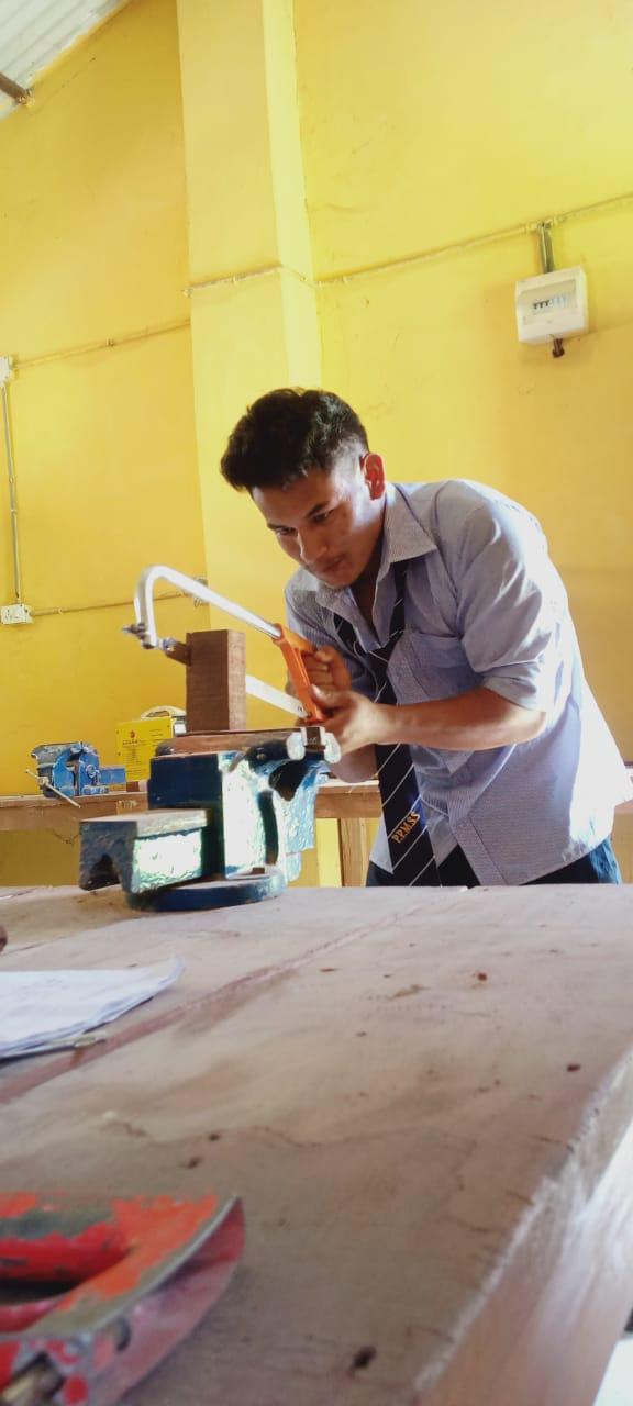 A person in a blue shirt and tie is using a hacksaw to cut wood secured in a vice on a workbench. The room has yellow walls, and various tools and papers are on the table.