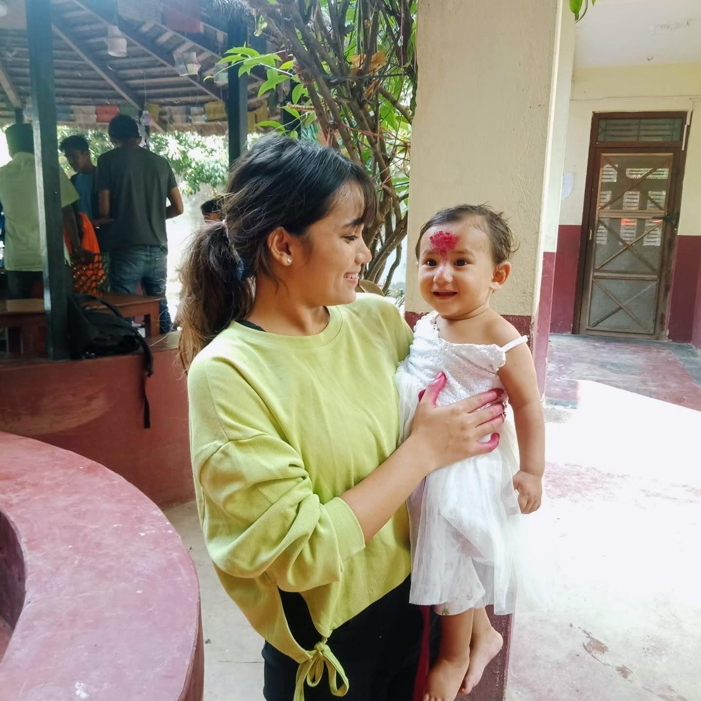 A woman in a green sweater is smiling and holding a child in a white dress with a red marking on the forehead. They are standing in a room with red and cream walls. Other people are in the background, and there is a tree nearby.