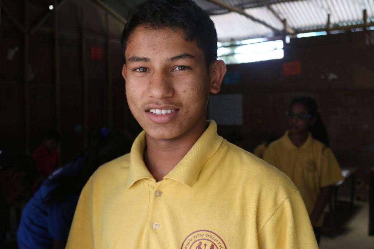 A young person wearing a yellow polo shirt smiles indoors. Other individuals in similar attire are visible in the dimly lit background. The setting appears to be a classroom with wooden walls and a corrugated metal roof.