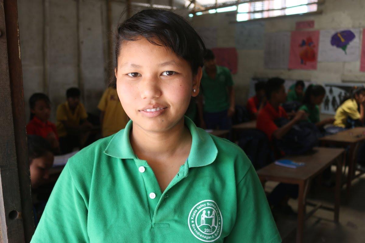 A young student wearing a green shirt stands smiling in a classroom. Other students are seated at desks in the background. The room has educational posters on the walls.