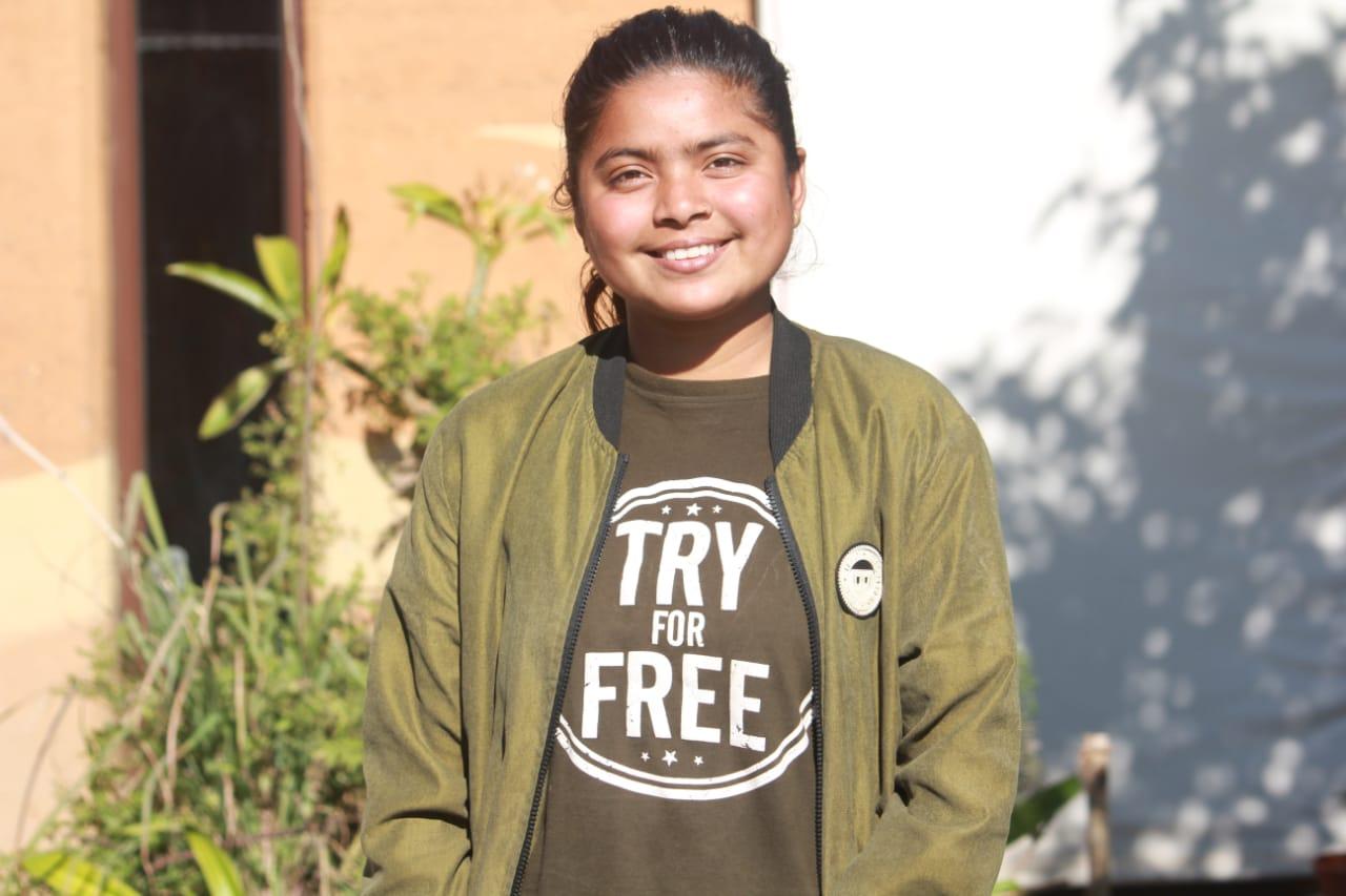 A person stands outside, smiling at the camera. They are wearing an olive-green jacket and a black t-shirt with the text "Try For Free." Green plants and a building's wall are visible in the background.