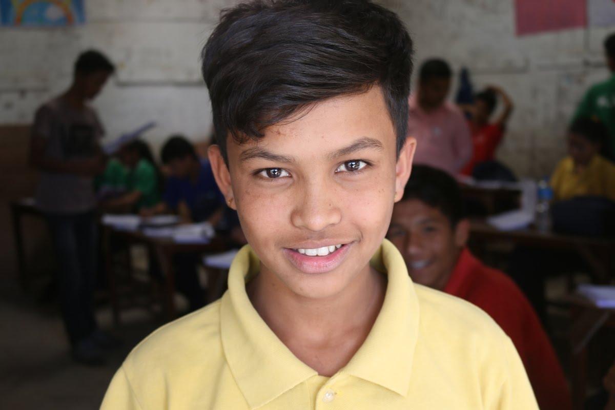 A boy wearing a yellow polo shirt smiles at the camera in a classroom setting. Other students sit at desks in the background, engaging with their work. The walls are adorned with educational posters.