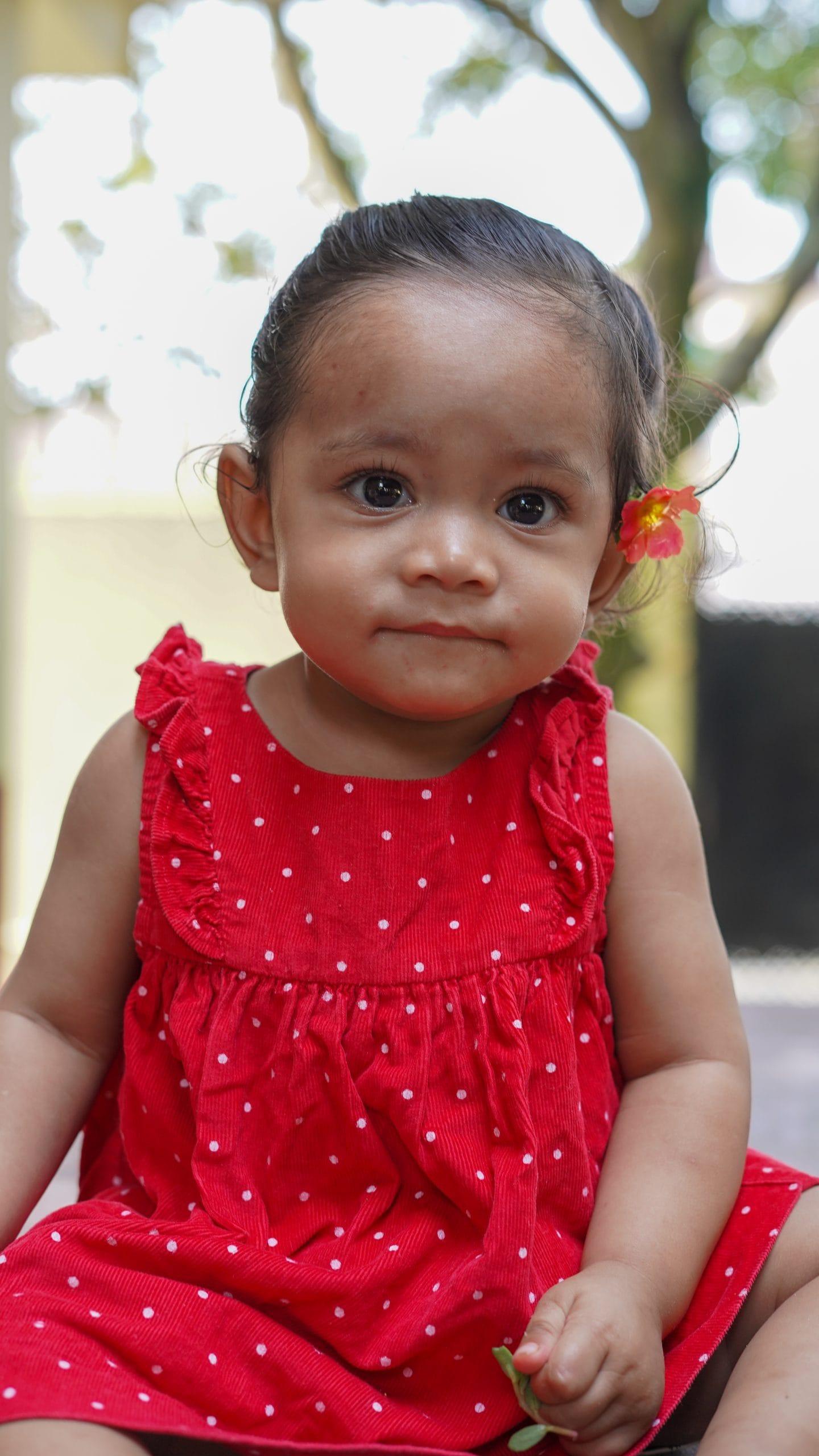 A toddler in a red polka dot dress sits outdoors, holding a small flower. She has a yellow and red flower tucked behind her ear, and there are blurred trees in the background.
