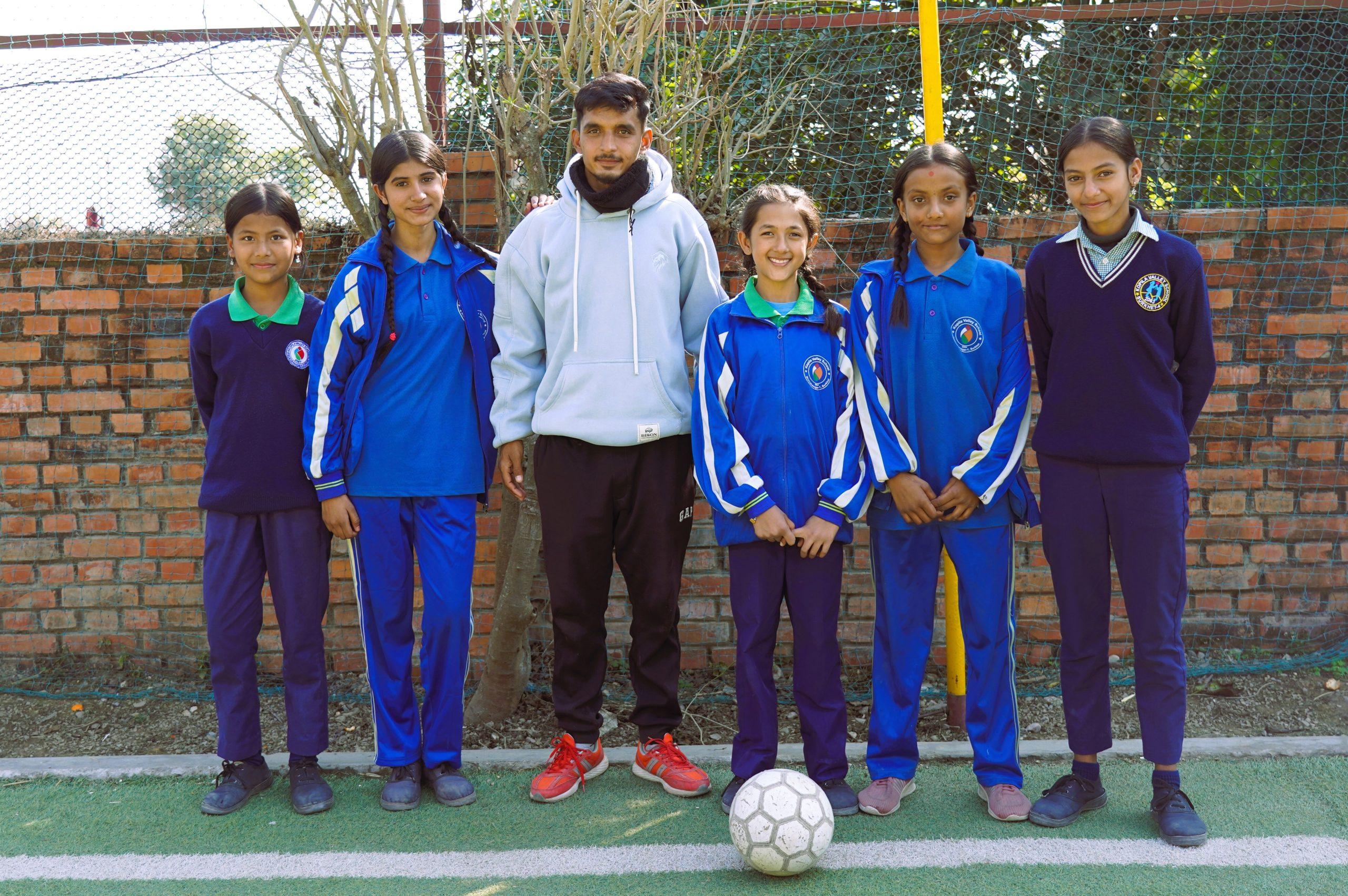 A coach in a gray hoodie and red sneakers stands with five young athletes in blue and navy sports uniforms. They are outdoors on a sports field with a soccer ball in front of them and a brick wall in the background.