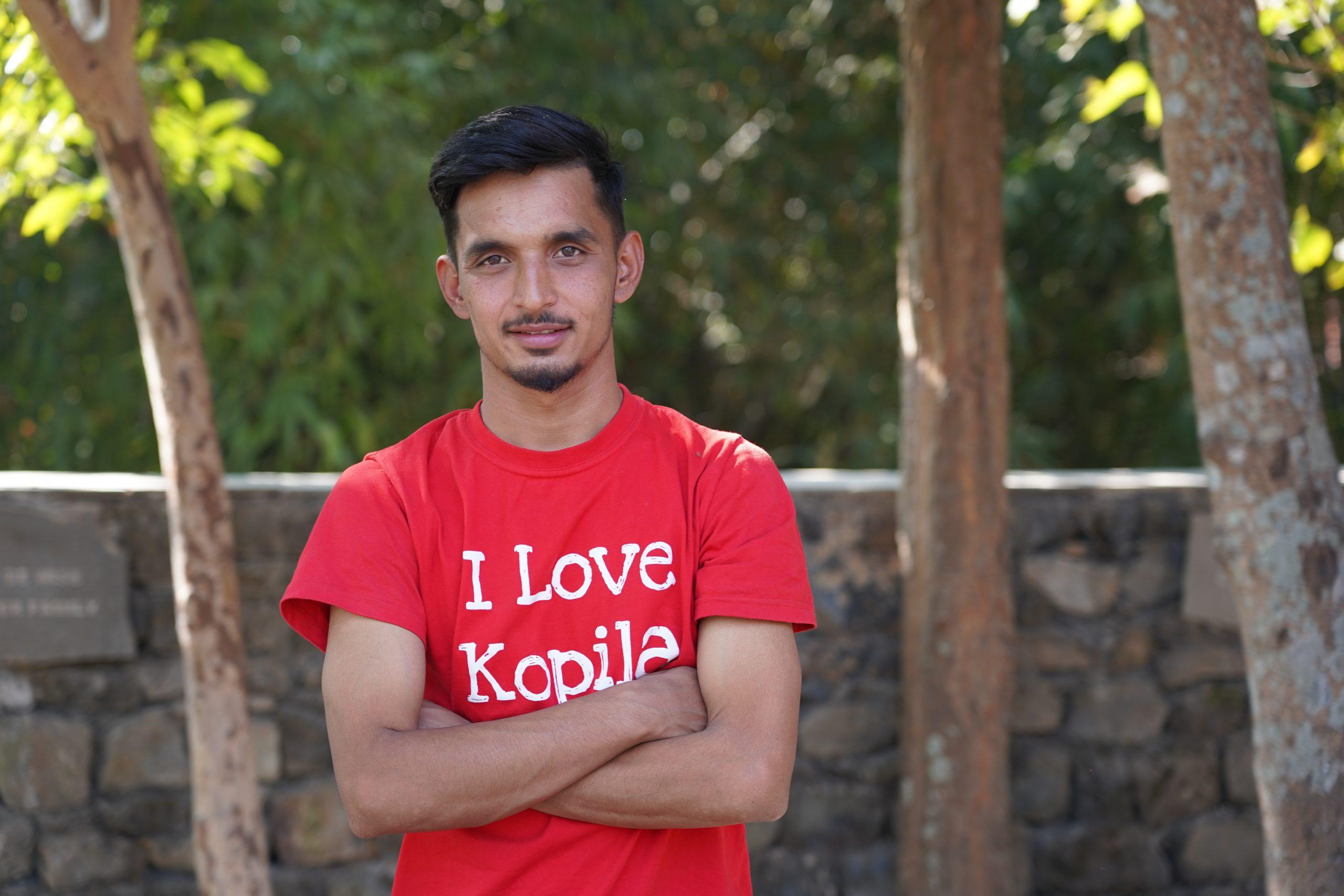 A man stands outdoors with arms crossed, wearing a red T-shirt that says "I Love Kopila." He is in front of a stone wall, with trees and greenery in the background.
