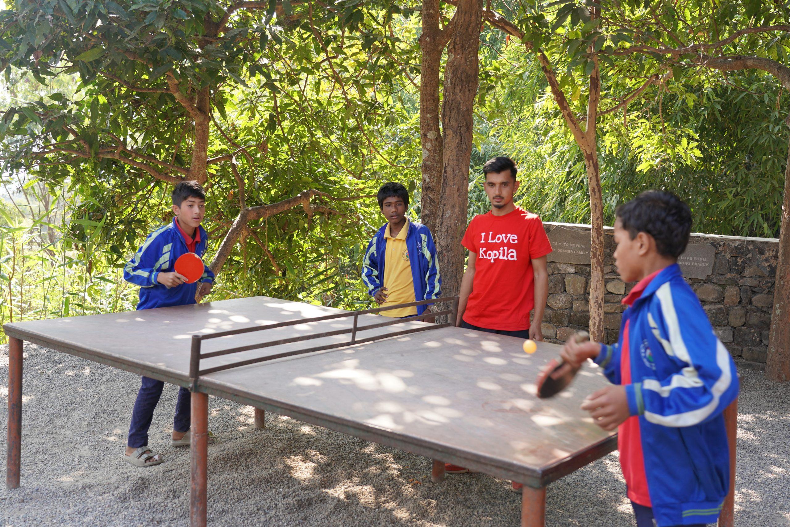 Four people are playing ping pong outdoors on a makeshift table. Two players hold paddles, while the others watch. A person in a red shirt with white text stands near a tree. The scene is shaded by trees.